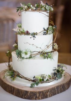 a three tiered white cake with greenery on top is sitting on a wood slice