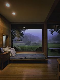 a bedroom with wooden floors and large windows overlooking the countryside at night, along with a bed that has been made up