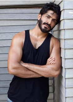 a man with his arms crossed leaning against a wall wearing a tank top and jeans