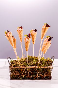 four ice cream cones with toppings on sticks in a basket filled with grass and dirt