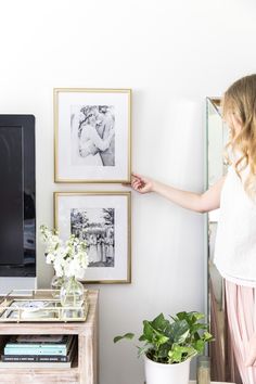 a woman pointing at pictures on the wall in her living room, with a television and potted plant