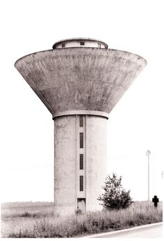 an old water tower sitting on the side of a road next to a tree and grass field