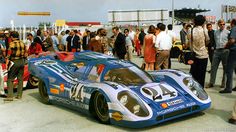 a group of people standing around a blue race car in front of some other people