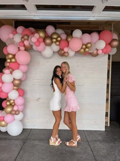 two women standing next to each other in front of a wall with pink and gold balloons