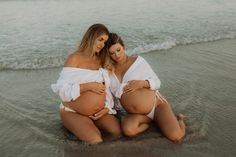 two pregnant women sitting on the beach with their backs to each other, both wearing white