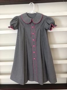 This child's size 6 dress was made with cotton gingham and it has 7 pink snaps on the front. It has a pleat on either side of the front band and two pleats in the back. It has a round collar and puffed sleeves with elastic that gathers the sleeve. There is pink mini-piping down the center band, around the collar, and on the edges of the sleeves. It measures 26' long from the highest point of the shoulder to the bottom of the hem. I can shorten this dress if needed. It can be washed in the washing machine and dried in the dryer. Pleated School Dress For Spring, Spring School Pleated Dress, Spring Pleated School Dress, Pleated School Dress For Summer, Pleated Summer School Dress, Summer School Pleated Dresses, Spring School Dress With Peter Pan Collar, Spring School Dress With Collar, Collared Spring Dresses For School