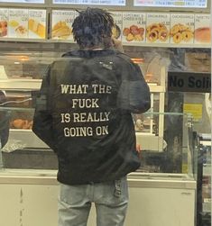 a man standing in front of a donut shop looking at his cell phone while wearing a jacket that says what the fock is really going on