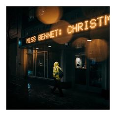 a man walking down the street in front of a building at night with lights on