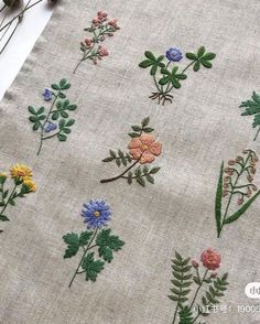 an embroidered piece of cloth with various flowers on it and some plants in the background