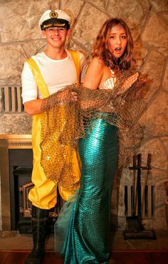 a man and woman dressed up as mermaids in front of a fire place with their arms around each other