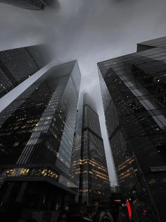 some very tall buildings in the city with lights on at night and dark clouds overhead