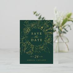 a green save the date card with gold foil on it and flowers in vase next to it