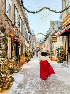 a woman walking down a snowy street with christmas decorations on the buildings and trees around her