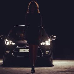 a woman standing in front of a car at night with the headlights on and behind her