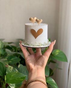 a hand holding a white cake with gold hearts on top and greenery in the background