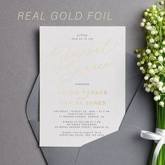 a white and gold foiled wedding card on top of a piece of paper next to a bouquet of baby's breath flowers