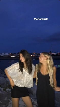 two beautiful young women standing next to each other near the water at night with city lights in the background