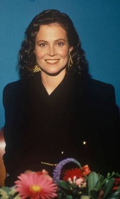 a woman sitting at a table with flowers in front of her and smiling for the camera