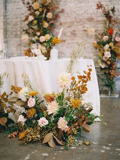 an arrangement of flowers and greenery on a table in front of a brick wall