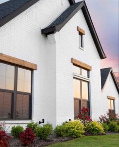 a white house with black shingles and red flowers in the front yard at sunset