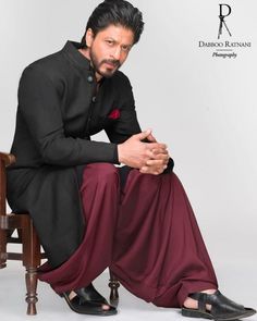 a man sitting on top of a wooden chair wearing a black shirt and maroon pants