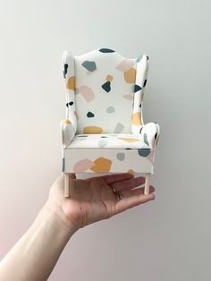 a hand holding up a miniature chair in front of a white wall with multicolored spots on it