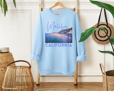 Get ready to ride the waves of style with our Malibu California sweatshirt! This amazing design features a captivating 3D-like graphic of Surfrider Beach at night, capturing the allure of this iconic surf spot. The word "Malibu" is elegantly written above in a playful and elegant script font, while "California" boldly stands below in a bold and stylish san-serif font, both in the same eye-catching color. Whether you're a beach lover, a surf enthusiast, or simply captivated by the coastal charm of Malibu, this sweatshirt is a must-have addition to your wardrobe. So, catch the cool California vibes and rock this stylish and cozy sweatshirt wherever your Malibu adventures take you! Designed with a perfect blend of soft cotton and breathable material, this classic heavy-blend crewneck sweatshi Beach Season Graphic Print Long Sleeve Sweatshirt, Long Sleeve Graphic Sweatshirt For Beach Season, Long Sleeve Graphic Print Sweatshirt For Beach Season, Beach Season Streetwear Sweatshirt Crew Neck, Relaxed Fit Sweatshirt For Beach Season Streetwear, Casual Sweatshirt With Front Print And Relaxed Fit, Blue Graphic Print Summer Sweatshirt, Blue Graphic Print Sweatshirt For Summer, Trendy Graphic Print Sweatshirt For Vacation