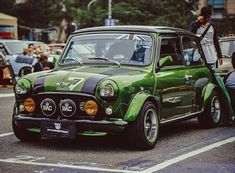 an old green car parked on the side of the road with people looking at it