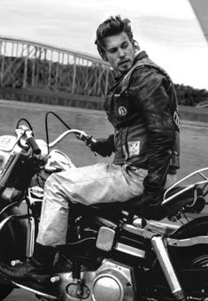 black and white photograph of a man sitting on a motorcycle with bridge in the background