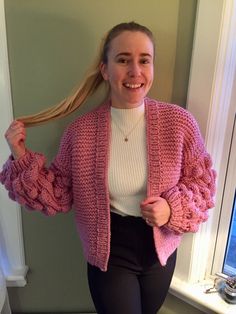 a woman standing in front of a window wearing a pink cardigan and black pants