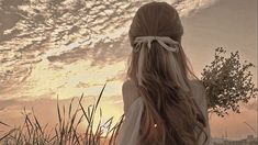 a woman with long hair standing in tall grass looking at the sun setting behind her