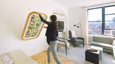 a woman standing in a living room holding up a large piece of artwork on the wall