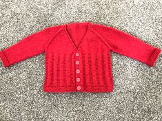 a red knitted cardigan sitting on top of a gray carpet next to a stuffed animal