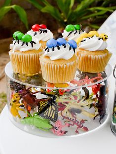 three cupcakes with white frosting and colorful decorations on a clear cake plate