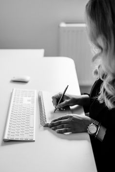 a woman sitting at a desk writing on a notepad with a laptop in the background