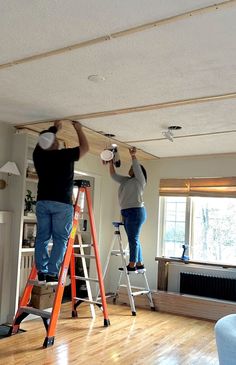 two people on ladders painting the ceiling in a living room with hard wood floors