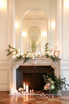 a fireplace with candles, flowers and greenery on it in a large room filled with white walls