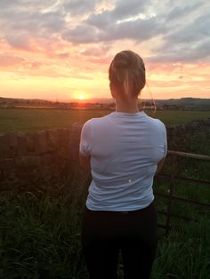 a woman standing in front of a fence watching the sun go down over a field