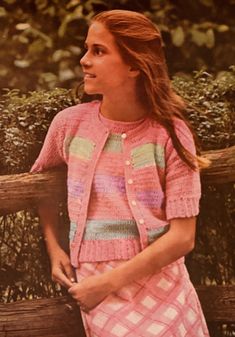 a woman standing next to a wooden bench in front of bushes and shrubbery wearing a pink cardigan