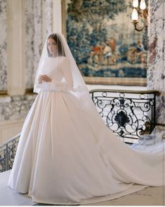 a woman in a white wedding gown and veil standing on a balcony with an ornate tapestry behind her