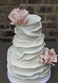 a three tiered white cake with pink flowers on top and brick wall in the background