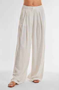 A person is wearing the Seychelles Relaxed Linen Pant in white, which features a loose-fitting, wide-leg design made from lightweight fabric. The pants epitomize relaxed style with front pleats, a high-rise elastic waistband, and a ground-length hem. Paired with flat, open-toed sandals, the outfit stands out against a plain white background.