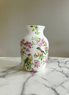 a white vase with pink flowers and birds on it sitting on a marble countertop