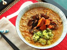 a bowl filled with noodles and meat on top of a red place mat next to chopsticks