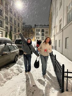 two people walking down a snowy street with cars parked on the side of the road