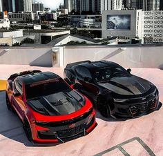 two black and red camaros parked on top of a roof with city in the background