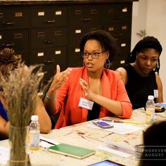 three women sitting at a table with papers and pens in front of them, talking to each other