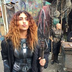 a woman with long hair holding a knife in front of graffiti covered walls and people