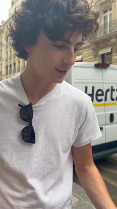 a young man with curly hair and sunglasses walking down the street in front of a bus