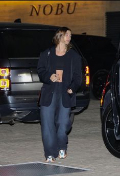 a woman in black jacket and pants walking next to a car with her hand on her hip